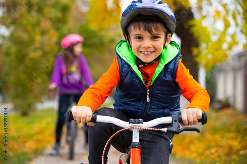 Portrait of a boy in the park with his sister in the background. Small child wearing a helmet and riding a bicycle on an autumn day. Active healthy outdoor sports