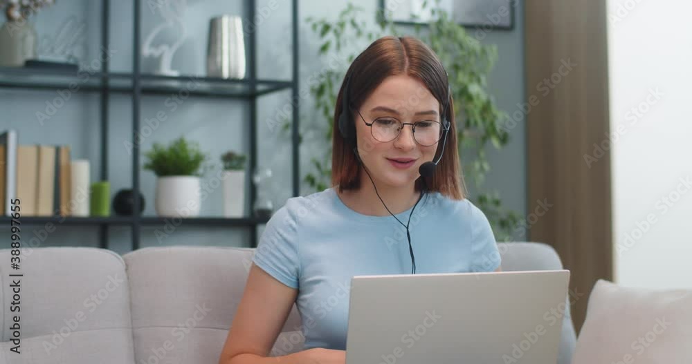 Charming Caucasian woman in glasses sitting on sofa and talking to friend via video conference. Smiling young female in headphones using laptop in living room. Communication, social media concept.