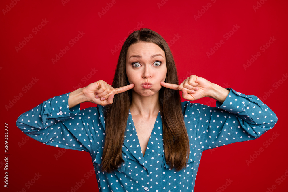 Close-up portrait of attractive funny playful girl pointing holding air in cheeks isolated on bright red color background Close-up portrait of attractive funny playful girl pointing holding air in cheeks isolated on bright red color background