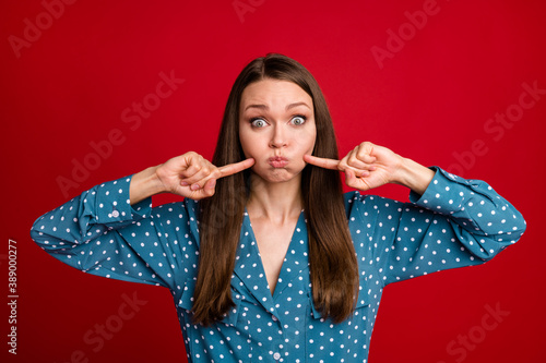 Close-up portrait of attractive funny playful girl pointing holding air in cheeks isolated on bright red color background