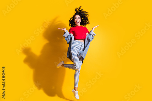 Full length photo of girl jump raise palms open mouth wear striped jeans cropped red t-shirt jacket sneakers isolated yellow color background