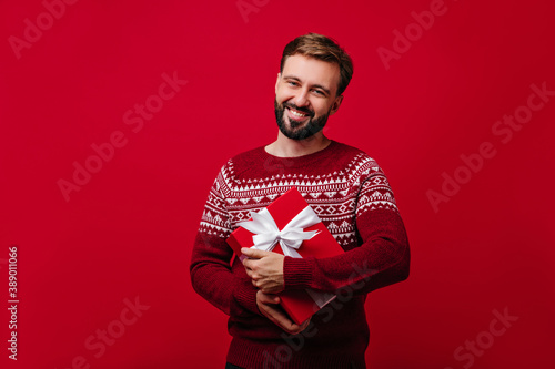 Pleased brunette man holding new year present. Indoor portrait of positive european guy smiling on red background. © Look!