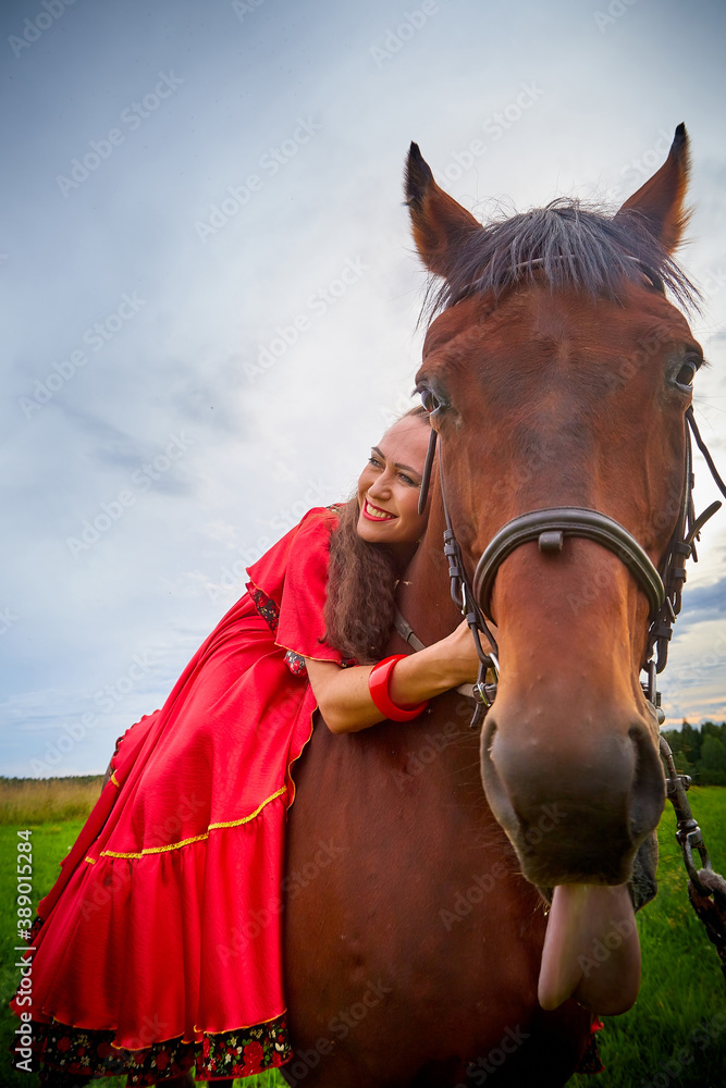 Obraz premium A woman in a bright Gypsy dress and image with a horse in a field with green grass. A model or actress posing in nature with an animal from a farm and the sky with clouds in the background