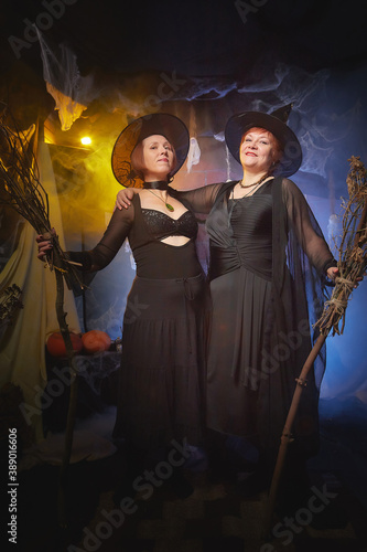 Two adult women looking as witches having fun and posing in dark room decorated for Halloween during photoshoot in studio.