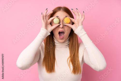 Young redhead woman over isolated pink background holding colorful French macarons with surprised expression