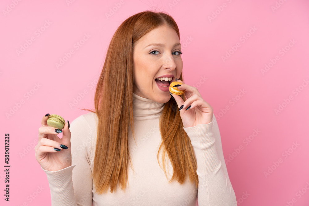 Young redhead woman over isolated pink background holding colorful French macarons and eating it Young redhead woman over isolated pink background holding colorful French macarons and eating it