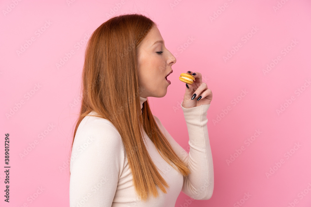 Young redhead woman over isolated pink background holding colorful French macarons and eating it Young redhead woman over isolated pink background holding colorful French macarons and eating it