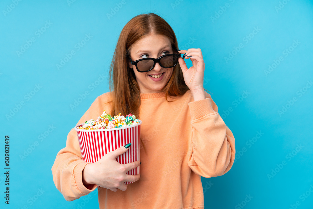 Obraz premium Young redhead woman over isolated background with 3d glasses and holding a big bucket of popcorns