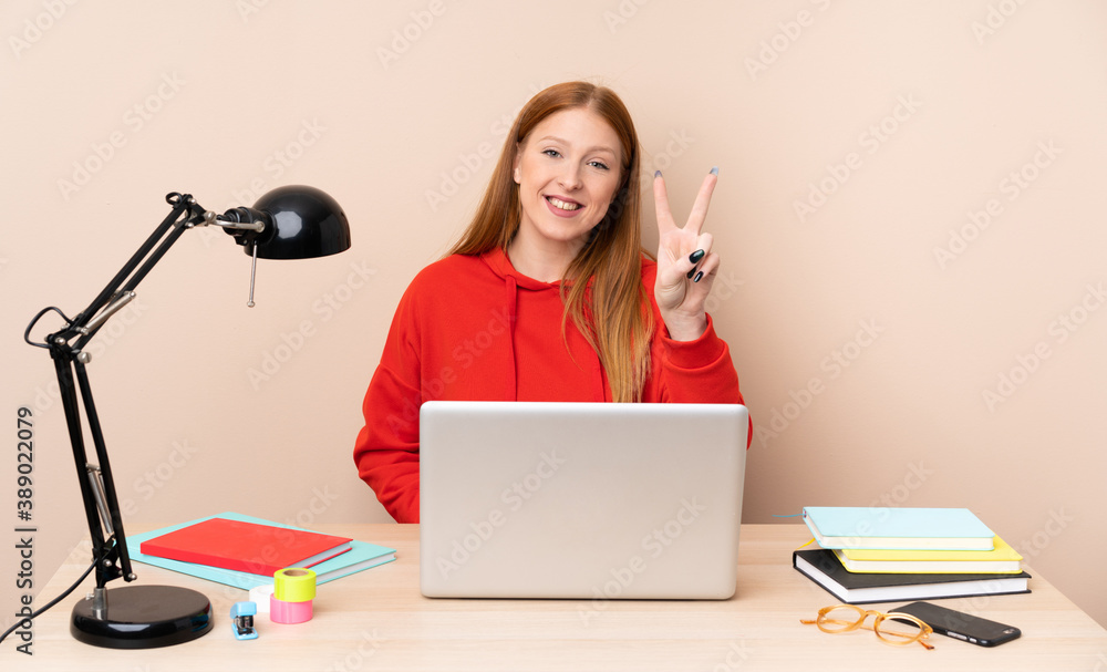 Young student woman in a workplace with a laptop smiling and showing victory sign Young student woman in a workplace with a laptop smiling and showing victory sign