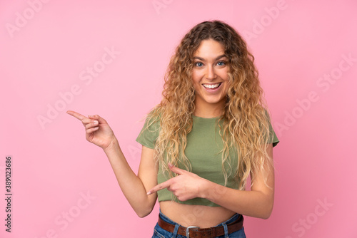 Young blonde woman with curly hair isolated on pink background surprised and pointing side