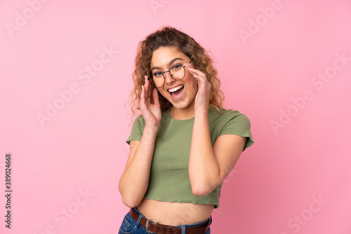 Young blonde woman with curly hair isolated on pink background with glasses and surprised