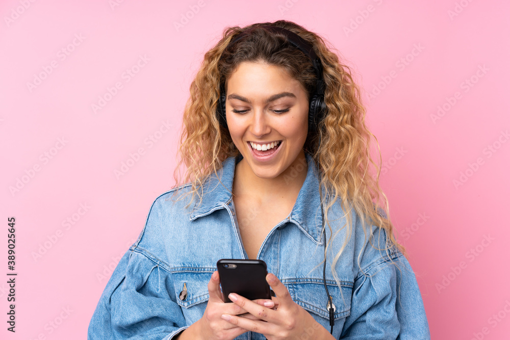 Young blonde woman with curly hair isolated on pink background listening music and looking to mobile Young blonde woman with curly hair isolated on pink background listening music and looking to mobile