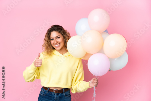 Young blonde woman with curly hair catching many balloons isolated on pink background giving a thumbs up gesture