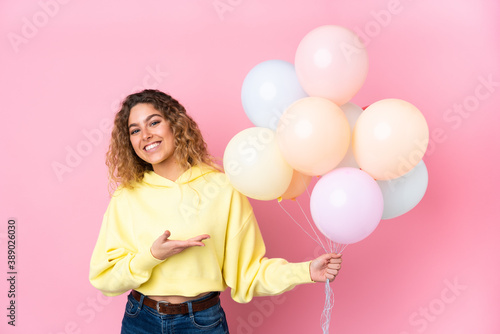 Young blonde woman with curly hair catching many balloons isolated on pink background extending hands to the side for inviting to come