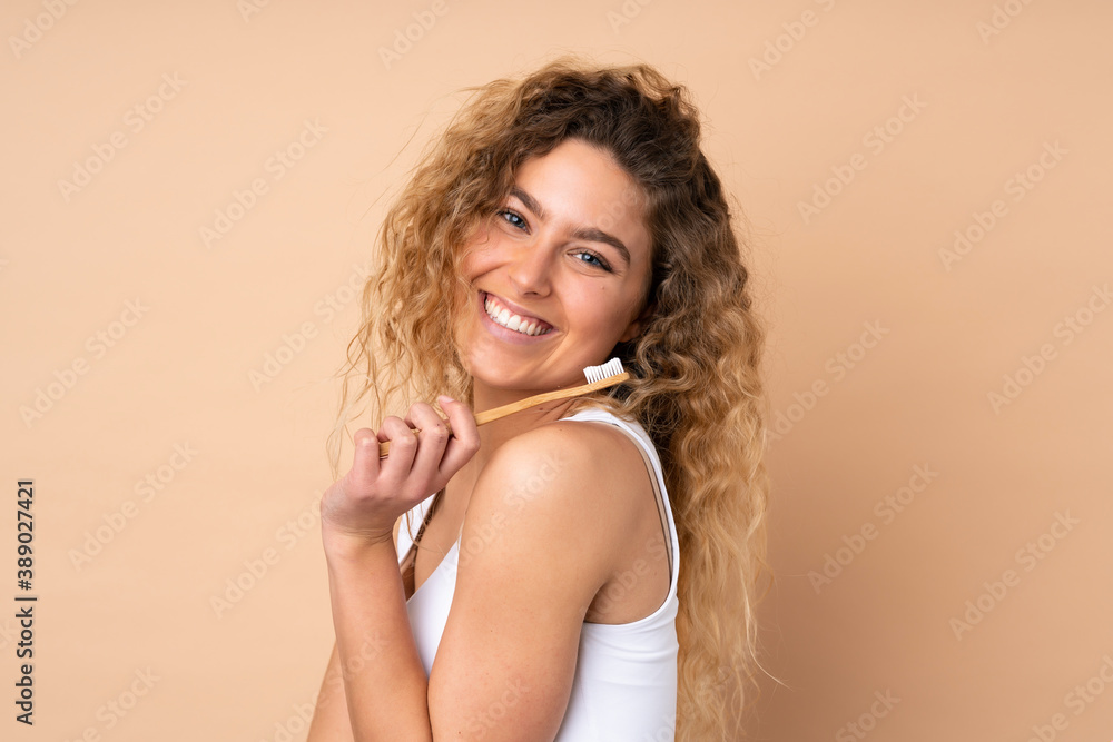 Young blonde woman with curly hair isolated on beige background with a toothbrush and happy expression Young blonde woman with curly hair isolated on beige background with a toothbrush and happy expression