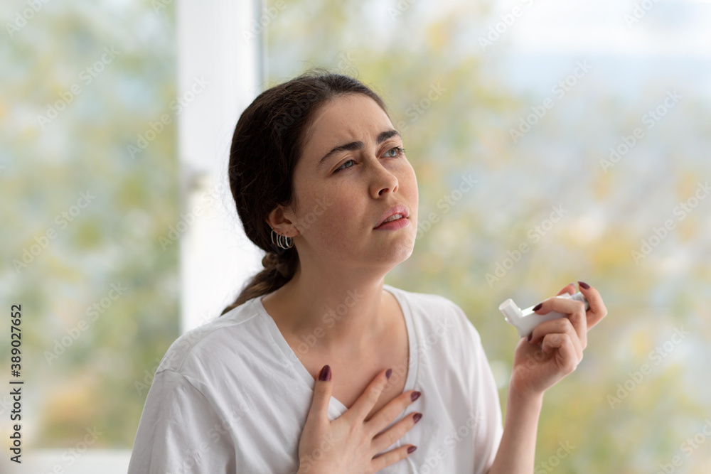 Allergic Asthma. Portrait of a woman holding an inhaler with medicine in her hand and clutching her chest, experiencing a spasm. Blurry background