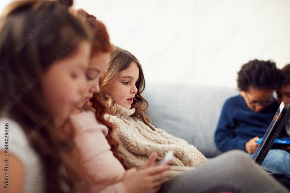 Group Of Children With Friends Sitting On Sofa At Home Playing Together On Digital Devices