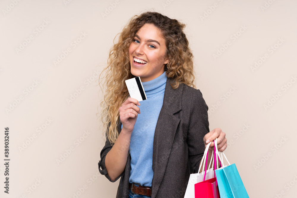 Young blonde woman with curly hair isolated on beige background holding shopping bags and a credit card Young blonde woman with curly hair isolated on beige background holding shopping bags and a credit card