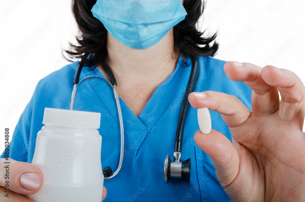 A female doctor in a blue uniform presents one white pill. She has a stethoscope around the neck. Protective mask on her face.