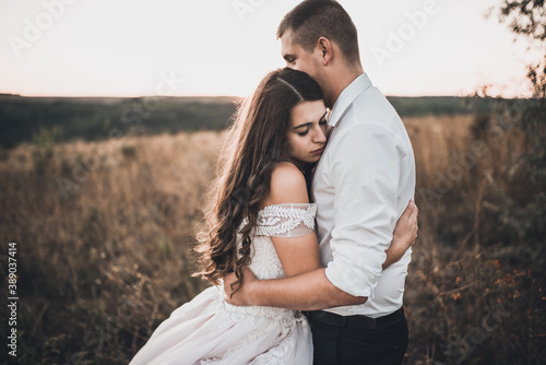 bride and groom in wedding white dress walking on the meadow in summer at sunset