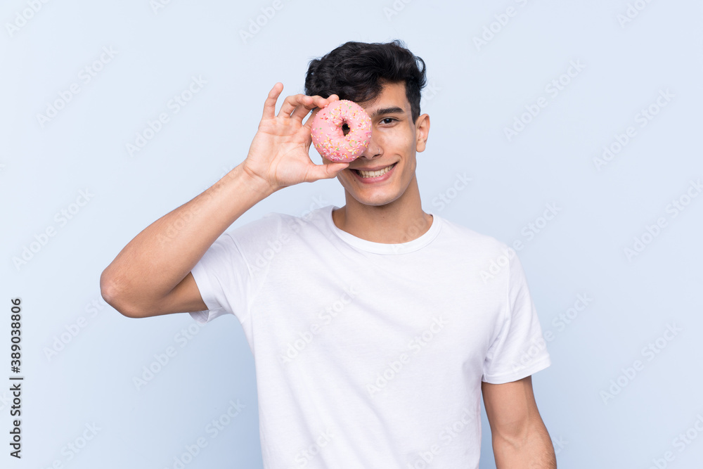 Young Argentinian man over isolated blue background holding a donut and happy Young Argentinian man over isolated blue background holding a donut and happy