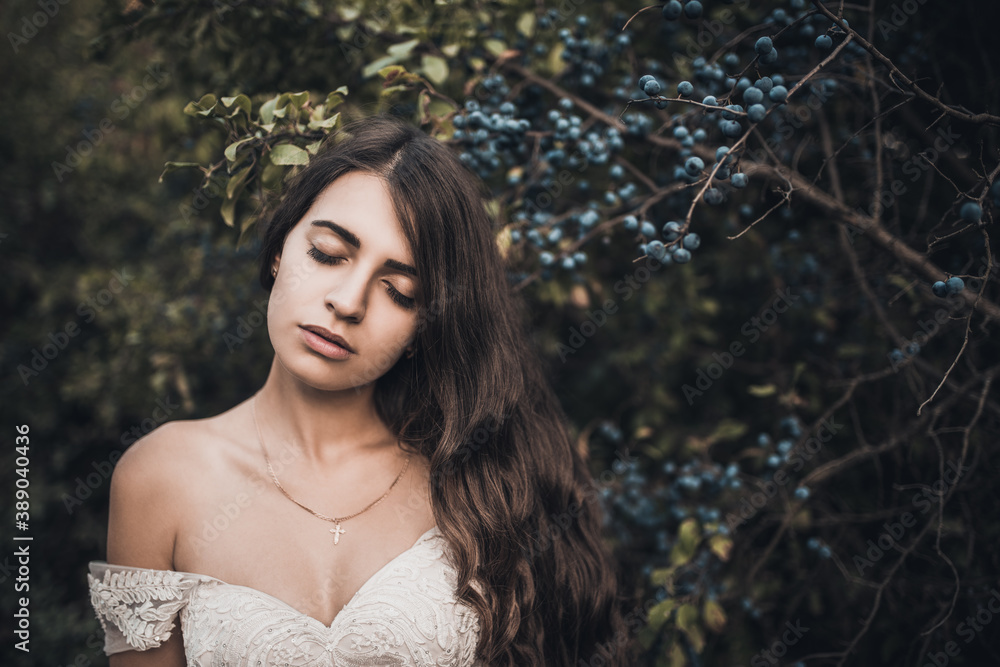 Woman Long-haired bride with bare shoulders near green bush with blueberries in summer Woman Long-haired bride with bare shoulders near green bush with blueberries in summer