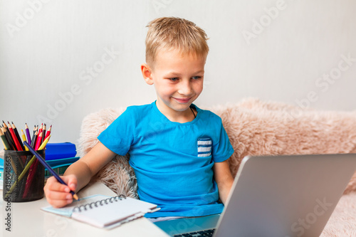 Little young school boy working at home with a laptop and class notes studying in a virtual class.