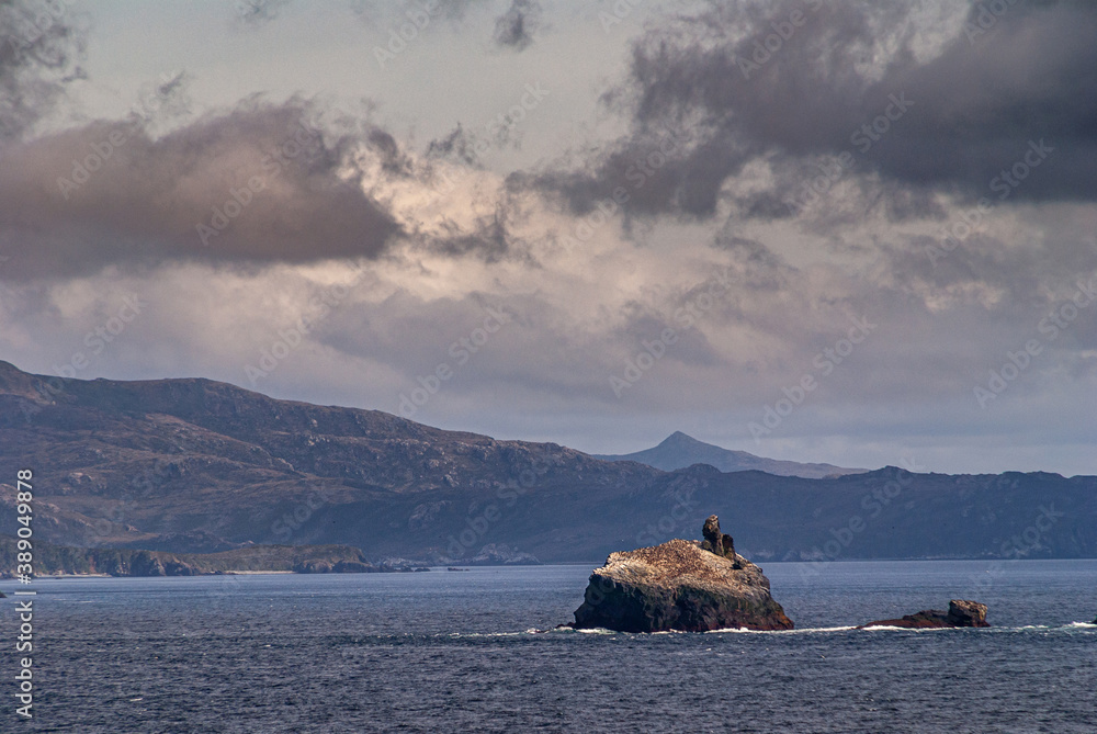 Cape Horn, Wollaston Islands, Chile - December 14, 2008: Closeup of rock Island soiled by birds in blue water under cloudscape. Larger island as backdrop.