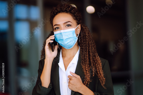 Beautiful serious business woman in protective medical mask standing near office and talking on phone. Safety during coronavirus pandemic, epidemic covid-19. 