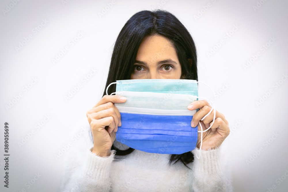 Brunette woman holds three surgical face masks close to face covering mouth isolated in grey background. Blue; green and white mask. Fashion and variety of corona accessories. Brunette woman holds three surgical face masks close to face covering mouth isolated in grey background. Blue; green and white mask. Fashion and variety of corona accessories.