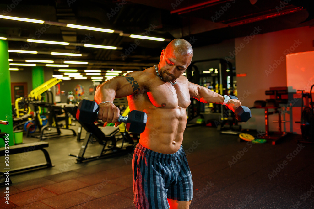 A strong, adult, fit, muscular male trainer poses for a photo shoot in a sports hall under the spotlights in sportswear, demonstrates his muscles and confidently holds dumbbells.