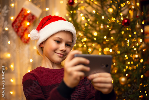 Cute young boy makes a selfie in the hat of Santa Claus, decorated fir-tree full of lights on the background. The concept of Christmas, holidays.