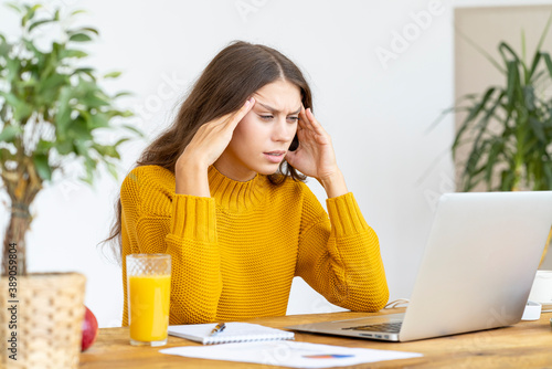 Woman massaging temples of head due to headache from long work at computer. Migraine. Beautiful young lady in bright yellow jumper is sitting at laptop