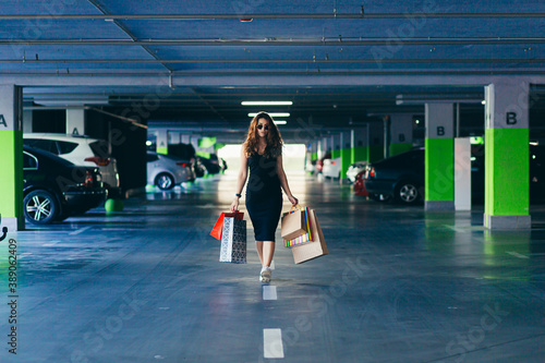 Cheerful lady in stylish black dress carrying on colorful shopping bags in a parking lot near a car. Girl after shopping. Female, caucasian. Attractive brunette looks satisfied after shopping at mall