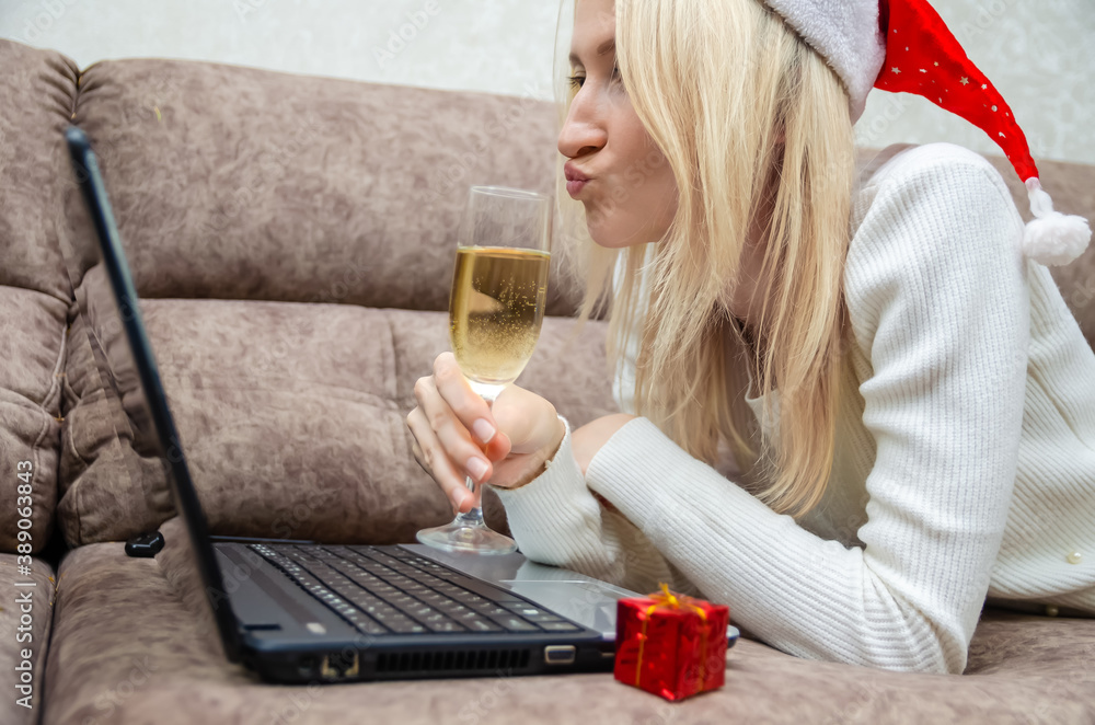Young beautiful blonde woman in a Santa hat is lying on the sofa with a laptop, a glass of champagne with a cheerful grimace on her face looking at the laptop camera. Concept of Christmas, New year. Young beautiful blonde woman in a Santa hat is lying on the sofa with a laptop, a glass of champagne with a cheerful grimace on her face looking at the laptop camera. Concept of Christmas, New year.