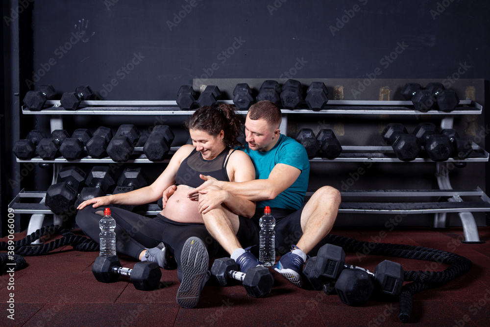 Beautiful sportive pregnant couple sitting on the floor hugging in the gym after a workout smiling and rejoicing. Fitness and healthy lifestyle during pregnancy. Family healthy lifestyle concept