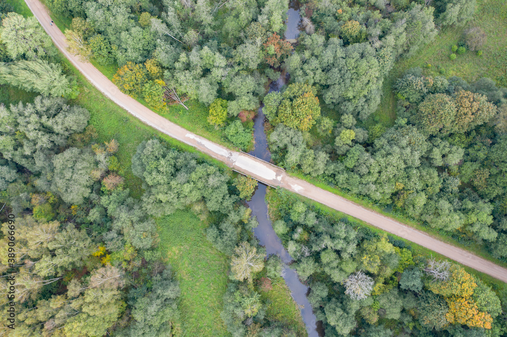 Aerial top down view of gravel road and bridge over winding river flowing through green forest
