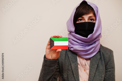 Portrait of young muslim woman wearing formal wear, protect face mask and hijab head scarf, hold Kuwait flag card against isolated background. Coronavirus country concept. photo