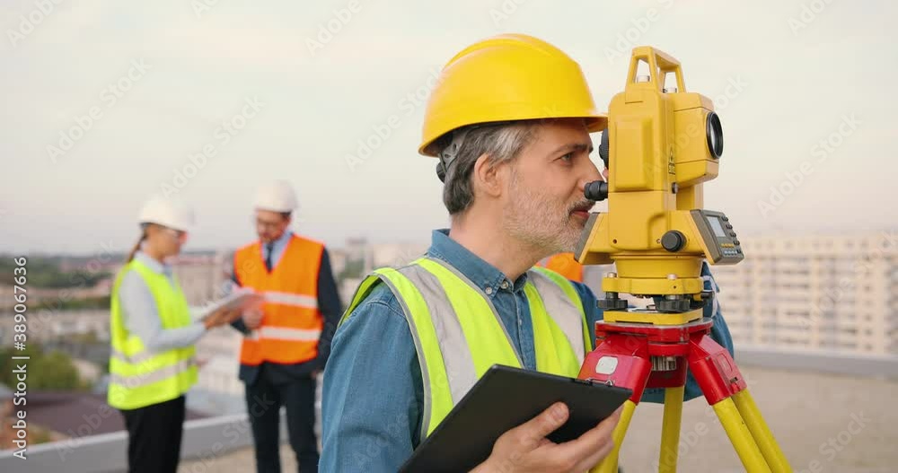 Caucasian man topographer in casque measuring angle with total station on building roof. Male builder constructor doing topographic measures and using tablet device. Geodesic concept. Device computer.