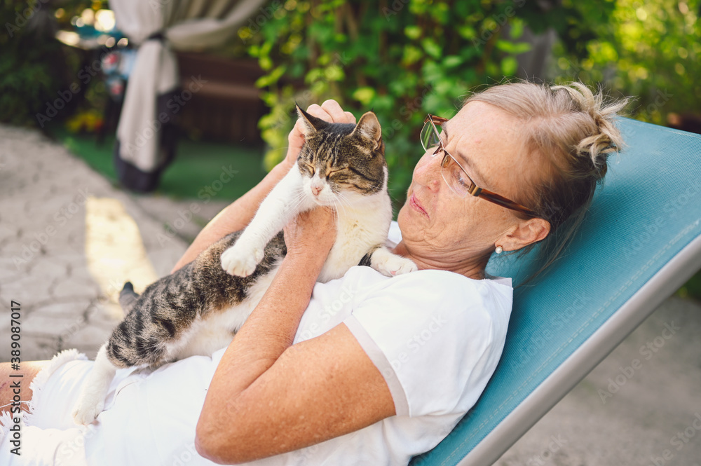 Happy smiling senior elderly woman in glasses relaxing in summer garden outdoors hugging domestic tabby cat. Retired old people and animals pets concept Happy smiling senior elderly woman in glasses relaxing in summer garden outdoors hugging domestic tabby cat. Retired old people and animals pets concept
