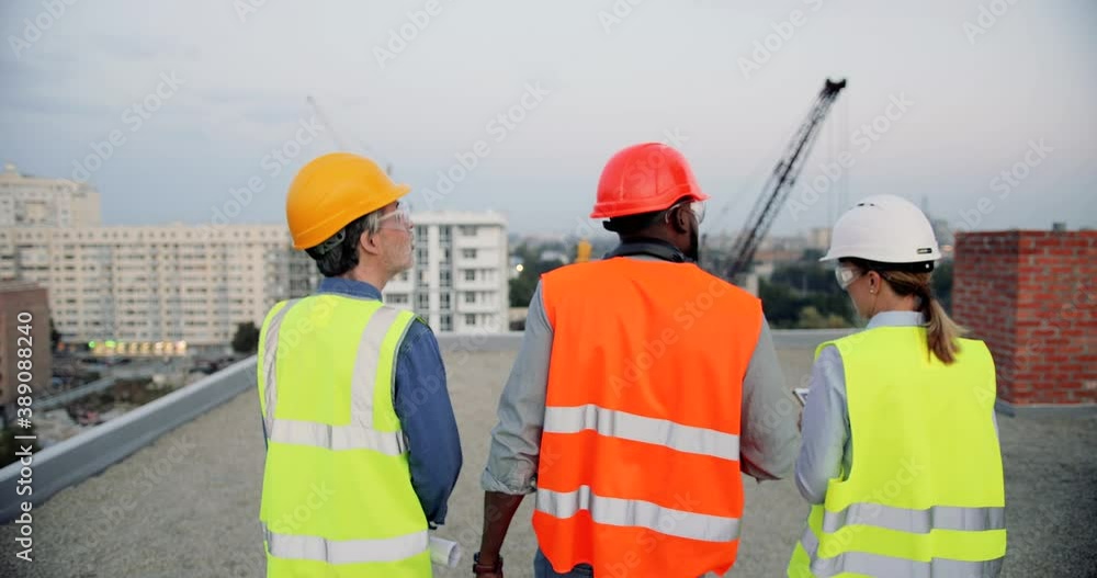 Rear. Mixed-races male and female builders in casques walking at roof top of building construction and talking. Back view on multi ethnic foreman and workers working and discussing constructing. Rear. Mixed-races male and female builders in casques walking at roof top of building construction and talking. Back view on multi ethnic foreman and workers working and discussing constructing.
