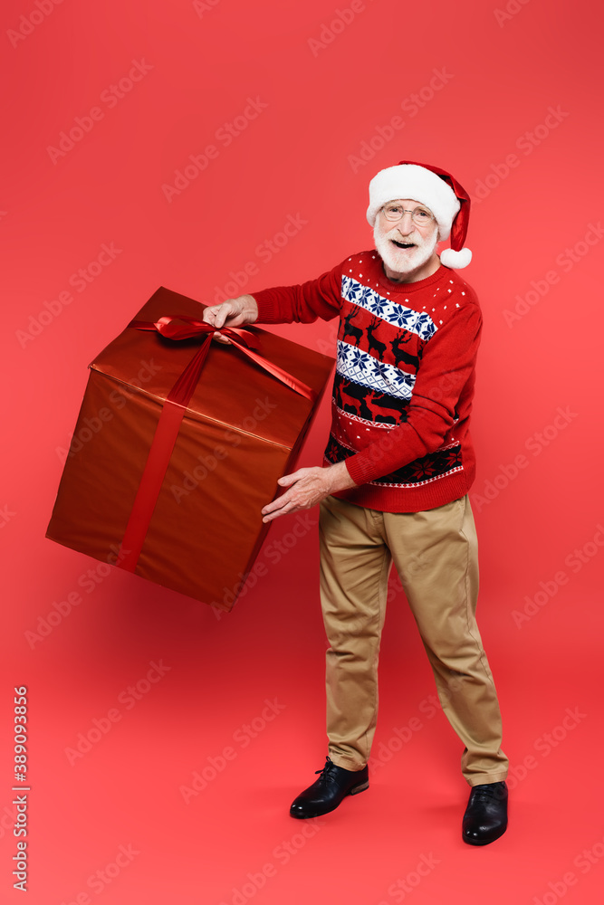 Smiling elderly man in santa hat holding present with bow and looking at camera on red background Smiling elderly man in santa hat holding present with bow and looking at camera on red background