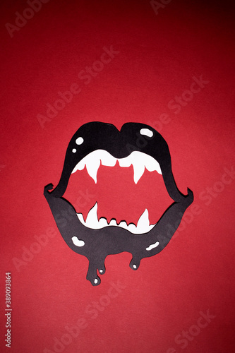 Black lips with white fangs cut out of paper on a red background. The vertical photo of paper art for your lovely scary Halloween design. There is a place for text.