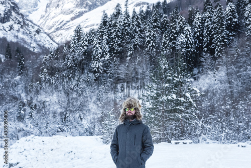 An adult brutal man with a beard in ski goggles and a hat stands in the mountains in winter. Active recreation concept