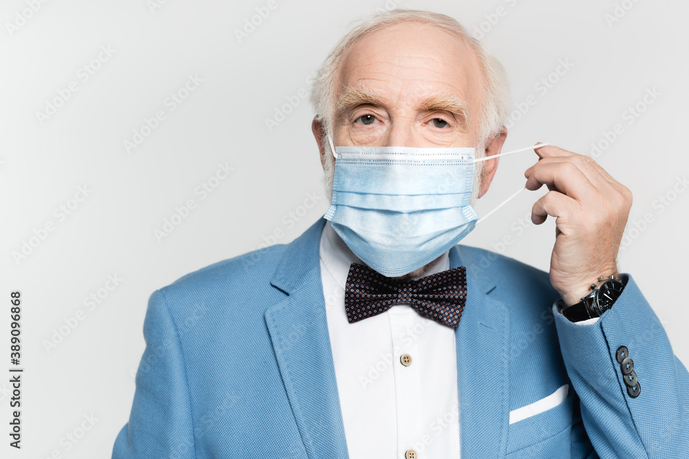 Elderly man in bow tie and jacket holding medical mask isolated on grey Elderly man in bow tie and jacket holding medical mask isolated on grey
