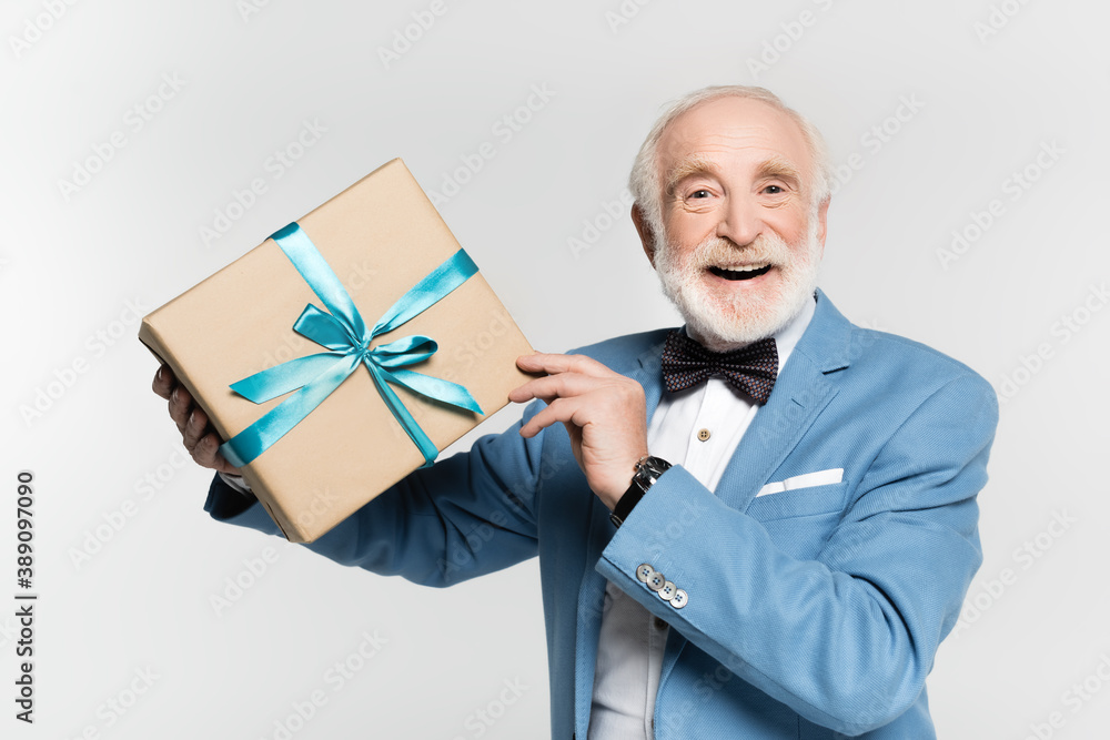 Smiling senior man in bow tie and jacket holding gift isolated on grey Smiling senior man in bow tie and jacket holding gift isolated on grey