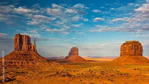 Dramatic western scene at monument valley with white clouds moving against a blue sky and red sandstone structures in the foreground