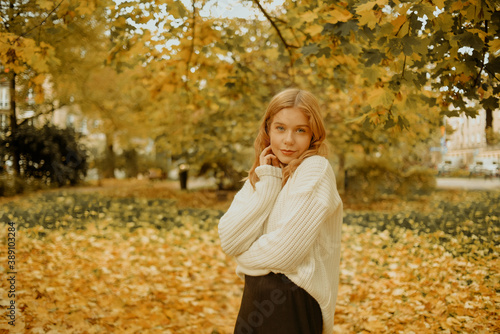 Woman in the autumn park. Girl teenager on a background of autumn leaves. Fashion photo of a model in a white warm sweater. Blonde looking at the camera