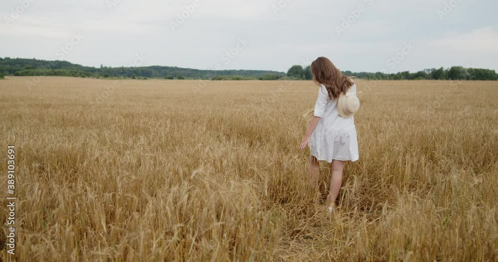 A young beautiful pregnant woman in a summer white dress with a hat behind her back and a wheat bouquet in a hand slowly walking through a wheat field, turning back, looking at the camera and smiling. A young beautiful pregnant woman in a summer white dress with a hat behind her back and a wheat bouquet in a hand slowly walking through a wheat field, turning back, looking at the camera and smiling.