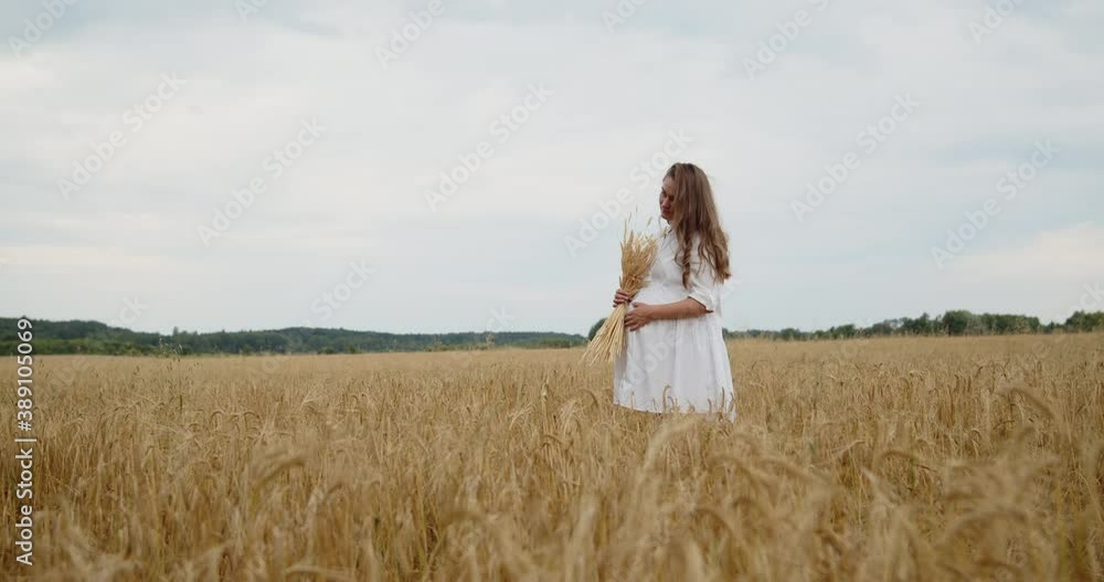 A young beautiful pregnant woman in a white summer dress stands in a wheat field holding wheat bouquet in hand and touching her belly A young beautiful pregnant woman in a white summer dress stands in a wheat field holding wheat bouquet in hand and touching her belly
